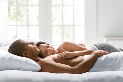 Sunlit bedroom with a rumpled dark gray comforter and white sheets on a bed, showing bare feet at the edge. Minimalist white headboard and wall in the background. Ideal for illustrating sleep comfort, temperature regulation, or morning routines