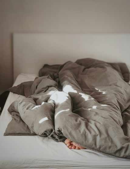Sunlit bedroom with a rumpled dark gray comforter and white sheets on a bed, showing bare feet at the edge. Minimalist white headboard and wall in the background. Ideal for illustrating sleep comfort, temperature regulation, or morning routines