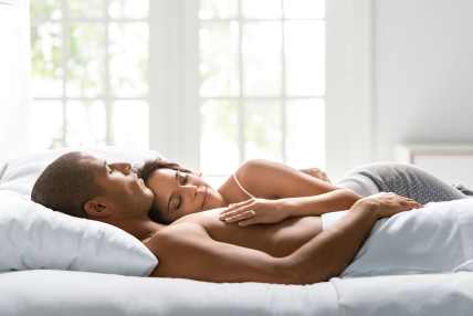 Sunlit bedroom with a rumpled dark gray comforter and white sheets on a bed, showing bare feet at the edge. Minimalist white headboard and wall in the background. Ideal for illustrating sleep comfort, temperature regulation, or morning routines