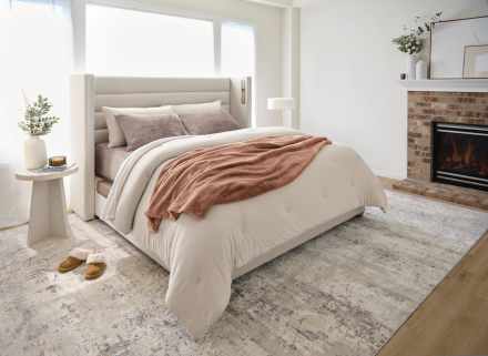 Warm and inviting bedroom featuring a beige upholstered bed with white bedding and a rust-colored throw blanket, set beside a cozy electric fireplace with decorative accents. The room includes a side table with a vase of greenery, candle, and book, plus brown slippers on a soft rug over hardwood floors, all bathed in natural light from large windows