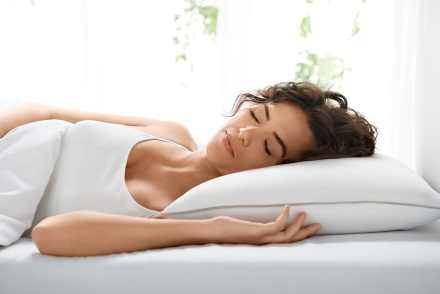 Neatly made bed with gray sheets and pillows, featuring a person lying down in soft focus, partially covered by the bedding. A bedside table with a small vase of pink flowers adds a touch of warmth to the serene sleep setting