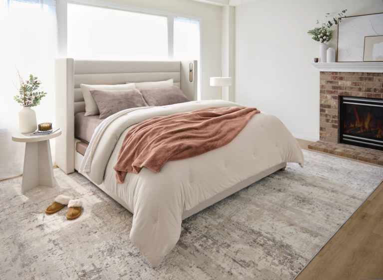 Warm and inviting bedroom featuring a beige upholstered bed with white bedding and a rust-colored throw blanket, set beside a cozy electric fireplace with decorative accents. The room includes a side table with a vase of greenery, candle, and book, plus brown slippers on a soft rug over hardwood floors, all bathed in natural light from large windows