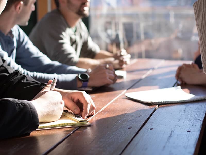 People seated at wooden table near window with notebooks and pens, contemplating something.