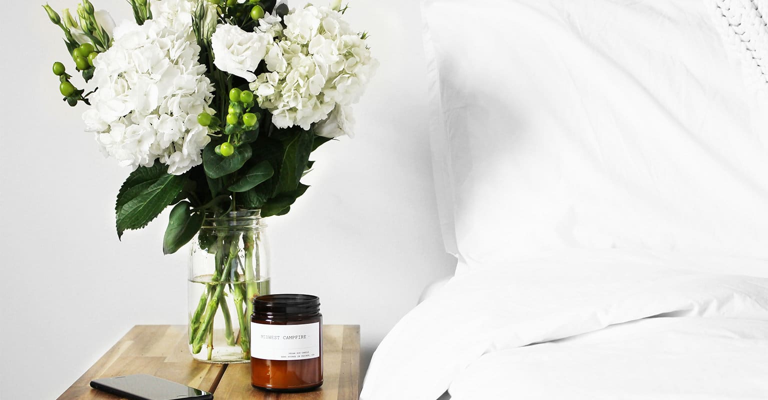 Bright white bedding and wooden bedside table with vase of white flowers, candle and cell phone.
