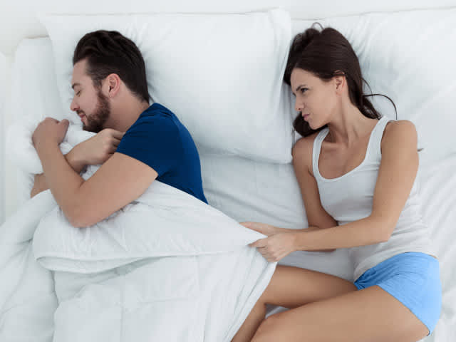 Couple sleeping in bed with man stealing the blanket
